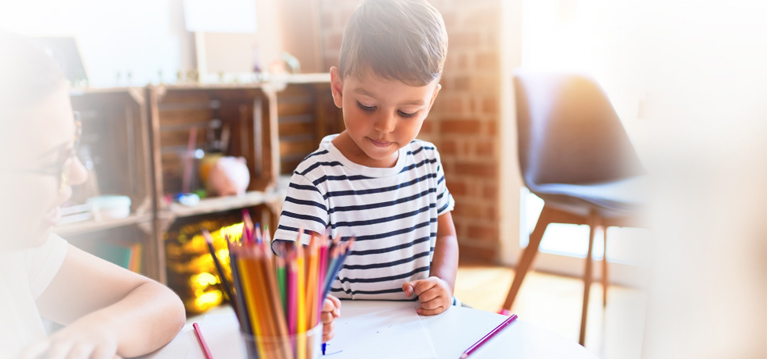 Beautiful teacher and toddler boy drawing draw using colored pencils at kindergarten