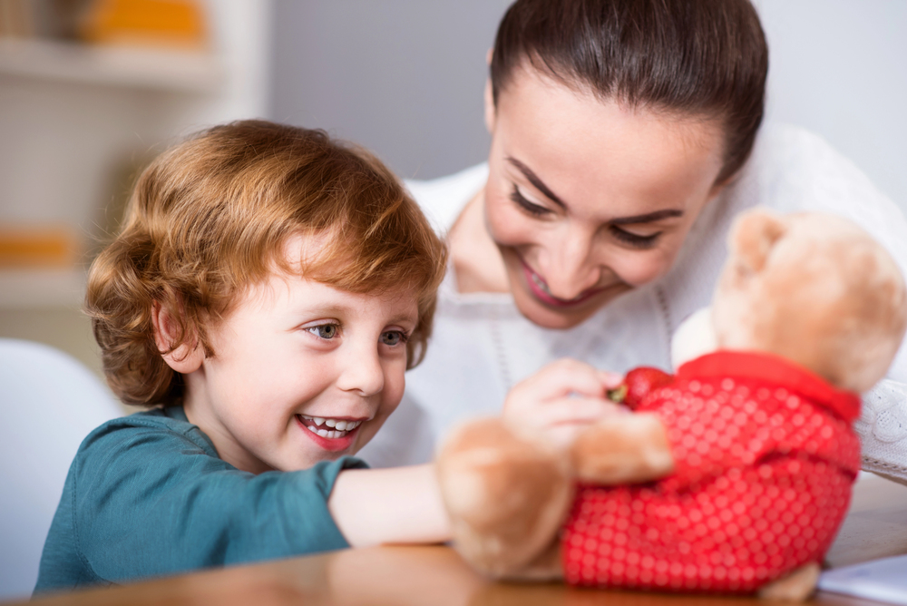 Smiling therapist engaging with young child and teddy bear.