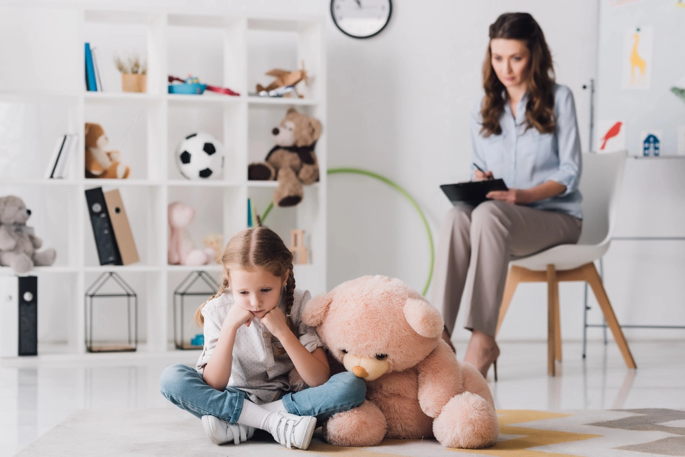 A young girl sitting on the floor with a sad expression, leaning against a large teddy bear, while a therapist takes notes in the background of a playroom filled with toys and shelves.