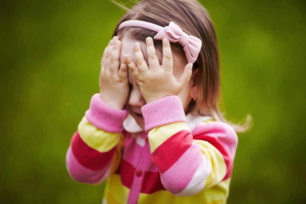 A young girl wearing a colorful striped sweater and a pink headband covers her face with her hands while standing outdoors on a grassy background.