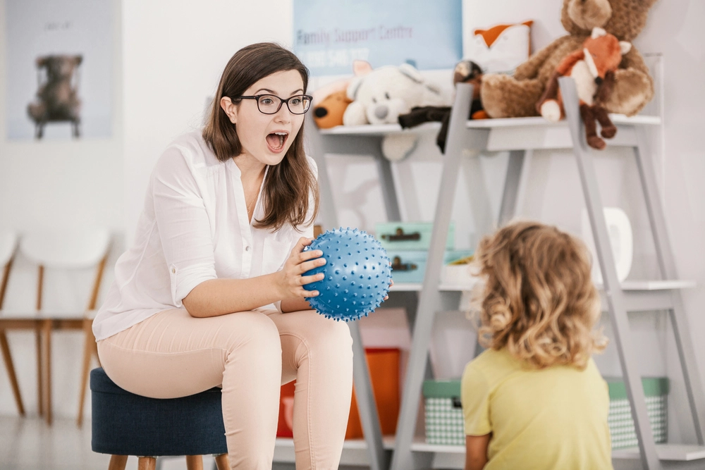 A female therapist in glasses and a white blouse enthusiastically interacting with a young child, holding a blue sensory ball in a playroom with toys and shelves in the background.