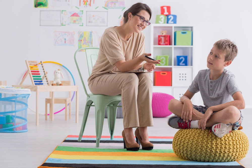 A female therapist sitting on a chair with a clipboard, engaging in a friendly conversation with a young boy seated on a yellow pouf in a colorful playroom filled with toys, drawings, and educational materials.